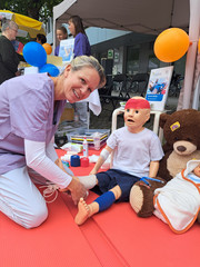 Eine Mitarbeiterin kniet am Boden mit einer Kinder-Übungspuppe und Stofftieren und blickt am Stand des Immanuel Klinikums Bernau Herzzentrum Brandburg beim Gesundheitstag in Bernau b. Berlin in die Kamera