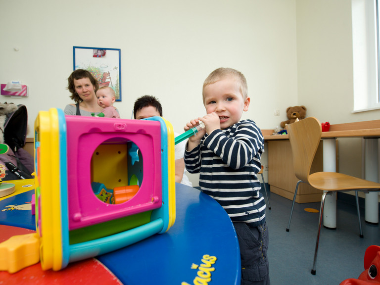 Ein Kleinkind steht in einem Untersuchungsraum mit einer Mutter mit Säugling und einem weiteren Kind, spielt mit buntem Spielzeug und blickt in die Kamera in der Kindermedizin des Immanuel Klinikums Bernau bei Berlin