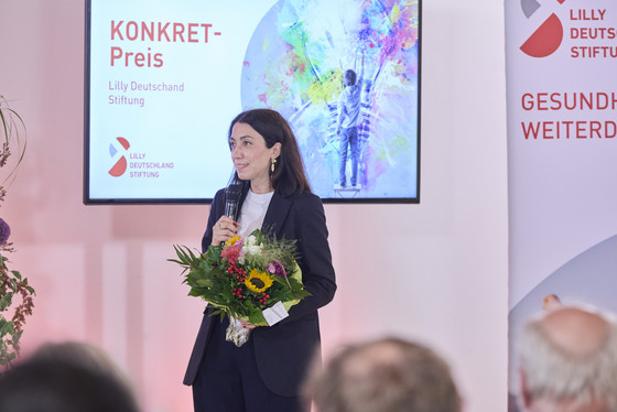 Dr. Viyan Sido steht mit Blumenstrauß auf der Bühne und spricht in ein Mikrofon